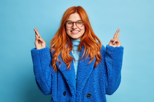 Portrait of happy woman with red natural hair smiles pleasantly keeps fingers crossed hopes for good luck dressed in winter fur coat transparent glasses.