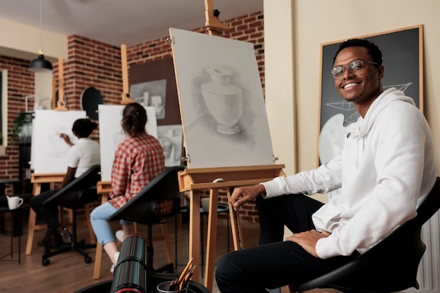 Free photo portrait of happy young african american man student of art school sitting at easel smiling at camera, learning pencil drawing in class. positive black guy enjoying creative hobby with friends