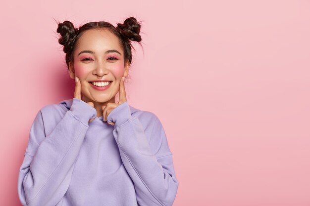 Портрет счастливой молодой азиатской девушки стоит в помещении, трогает щеки указательными пальцами, с приятной улыбкой на лице, одетая в повседневную фиолетовую толстовку с капюшоном, носит макияж в стиле кинозвезды.