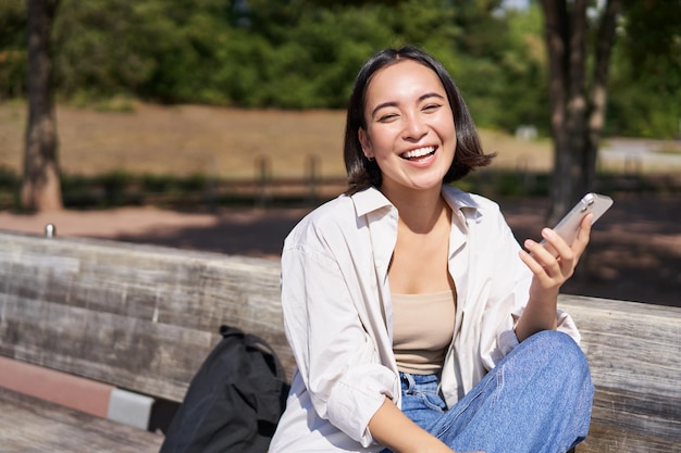 Portrait of happy young asian woman talking on mobile phone in park sitting on bench and having a te