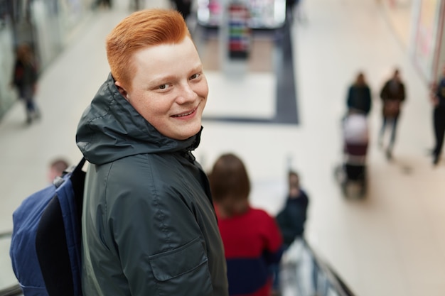 ショッピングモールに戻って立っている赤い髪の幸せな若い魅力的なヒップスターの肖像画