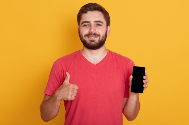 Портрет счастливого молодого бородатого хипстерского человека, носящего красную повседневную футболку и показывающего хорошо жест, держа телефон с пустым экраном