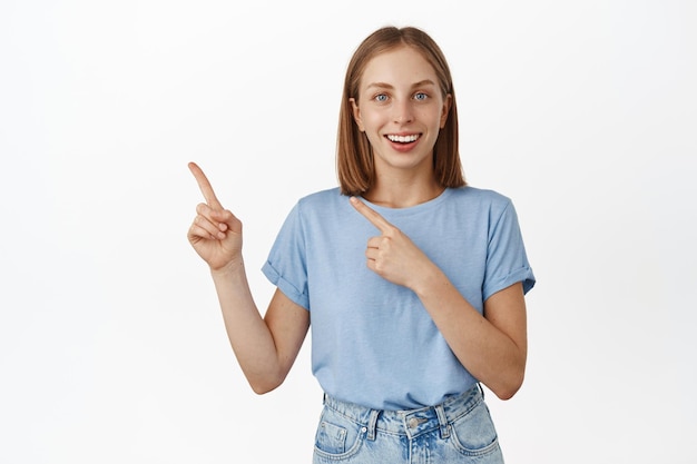 Портрет счастливой молодой блондинки в футболке, указывающей пальцем на распродажу, показывая рекламу в сторону, баннер со скидкой, логотип компании, рекомендую нажать на ссылку, белый фон.