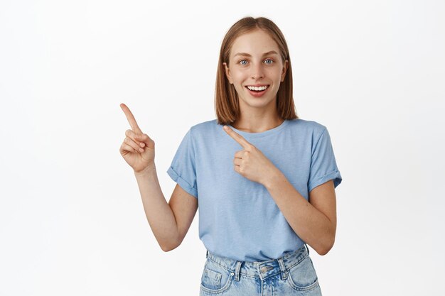 Portrait of happy young blond woman in t-shirt pointing fingers at sale, showing advertisement aside, discount banner, logo of company, recommend to click on link, white background.