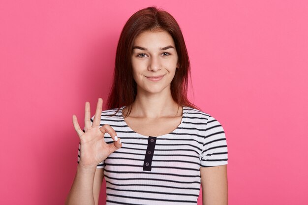 Portrait of happy young brunette girl showing ok gesture with fingers,  with charming smile, has excellent news, wearing striped t shirt.