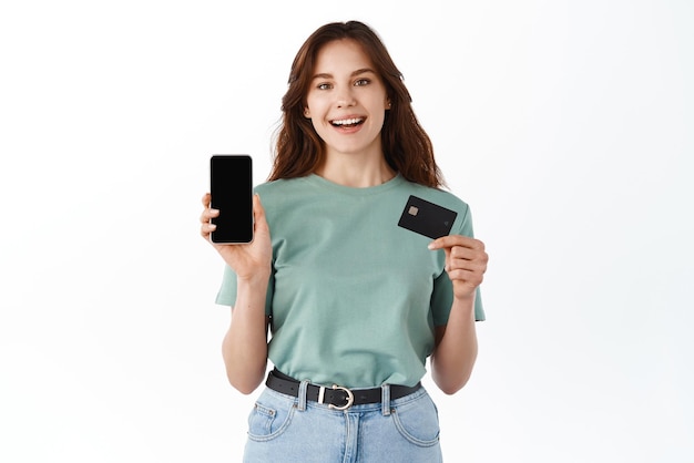 Portrait of a happy young brunette girl showing plastic credit card while holding mobile phone isolated over white background