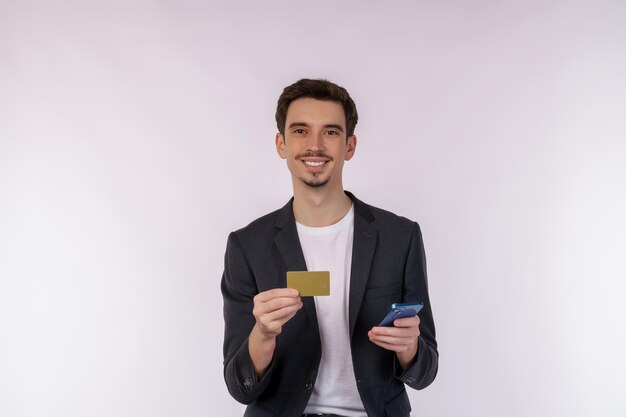 흰색 배경 스튜디오에서 격리된 신용 은행 카드를 들고 휴대 전화를 사용하여 서 있는 행복한 젊은 사업가의 초상화