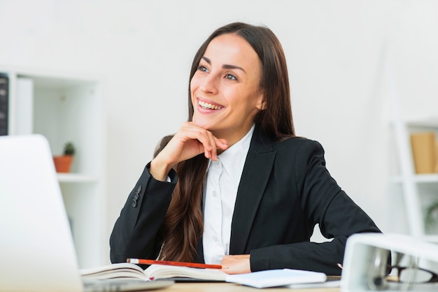 Free photo portrait of happy young businesswoman sitting at workplace daydreaming