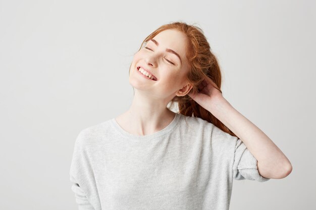 Portrait of happy young cute redhead girl smiling with closed eyes touching hair .