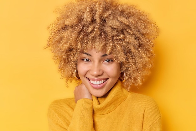 Free photo portrait of happy young delighted woman keeps hand on neck smiles pleasantly expresses sincere emotions keeps hand on neck wears warm jumper isolated over yellow background. human emotions concept