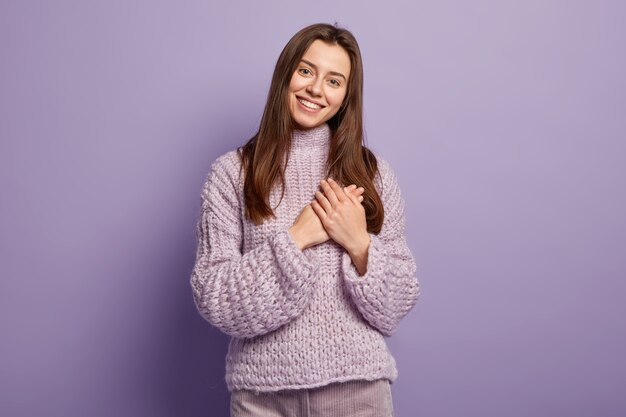 Portrait of happy young European woman keeps hands on breast, shows heart gesture, expresses gratitude, being thankful, models against purple wall Body language. Monochrome. People and devotion