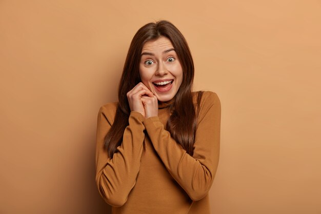 Portrait of happy young European woman looks pleased with admiration and joy , laughs as receieves great news, wears casual turtleneck, poses against beige wall. Emotions concept