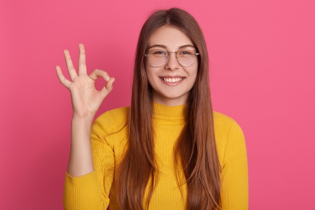 Portrait of happy young girl with long hair showing ok gesture with her fingers, looks smiling  being in good mood