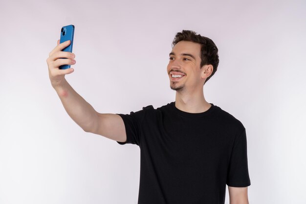 Портрет счастливого молодого красивого мужчины в черной футболке, держащего телефон и делающего селфи-фото на белом фоне