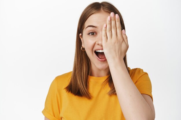 Portrait of happy young woman cover half of face, laughing and smiling amazed at front, stare surprised, standing in yellow t-shirt against white wall