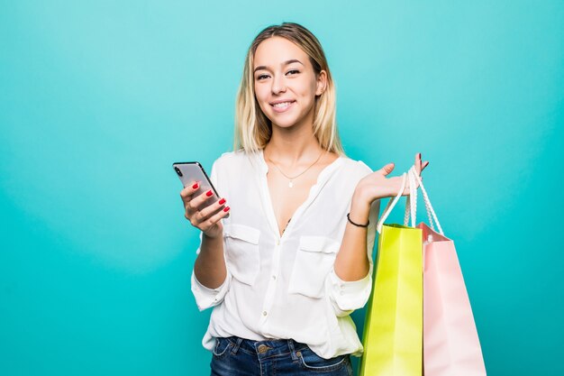 Portrait of a happy young woman holding shopping bags and mobile phone isolated on a mint wall