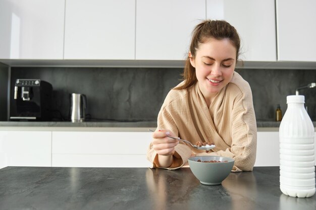 キッチンのワークトップにもたれかかり、シリアルを食べる幸せな若い女性の肖像画は、前にミルクとボウルを持っています