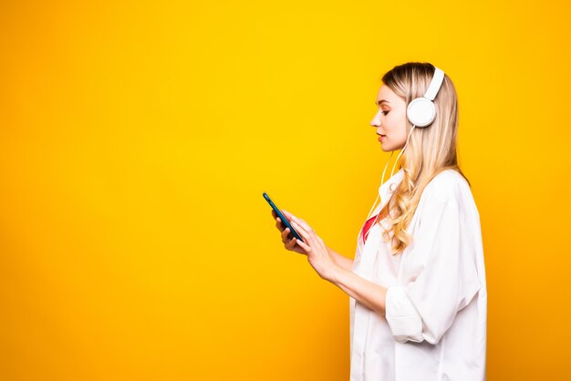 Portrait of a happy young woman listening to music with headphones and mobile phone isolated over yellow wall