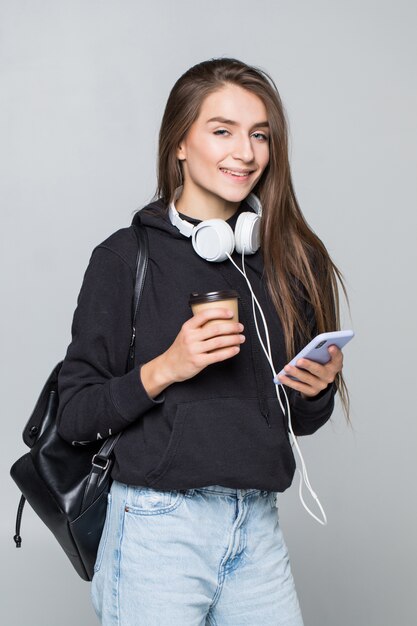 Portrait of happy young woman listening to music with mobile phone and earphones isolated on white wall