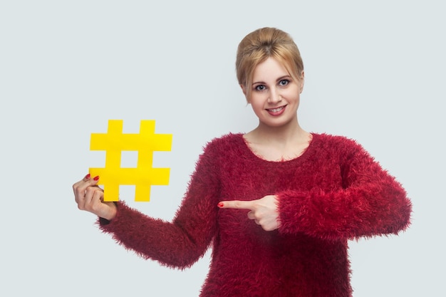 Portrait of happy young woman in red blouse standing and holding large big yellow hashtag sign and pointing finger social media concept indoor isolated studio shot copy space gray background