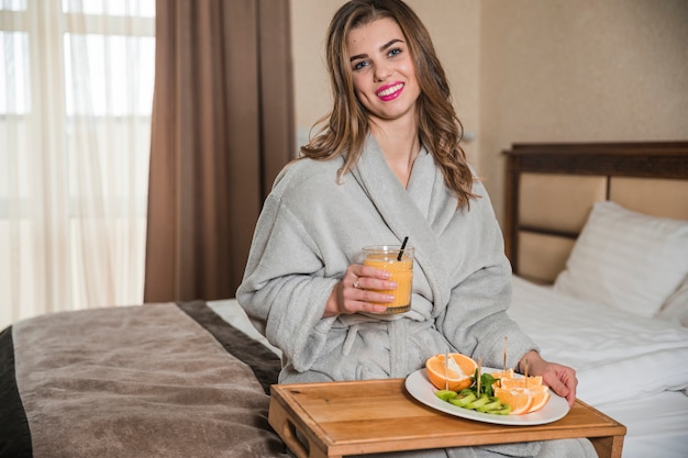 Portrait of a happy young woman sitting on bed holding glass of juice and healthy slices of fruits on plate