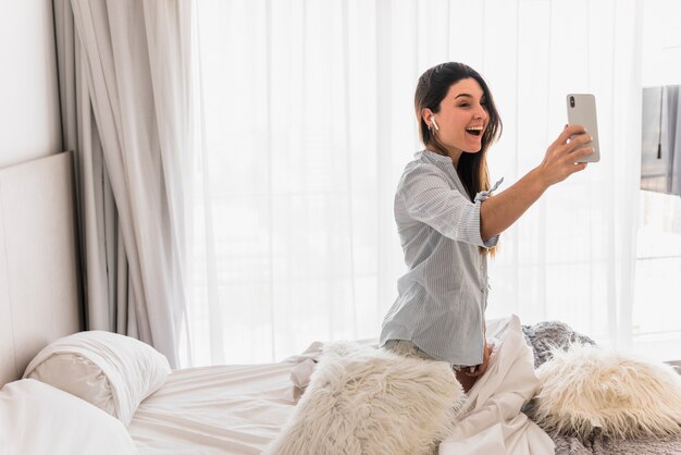 Portrait of a happy young woman sitting on bed taking making video call on mobile phone