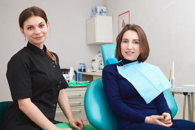 Portrait of happy young woman sitting in dental chair while visiting female dentist for routine checkup, teeth cleaning or whitening