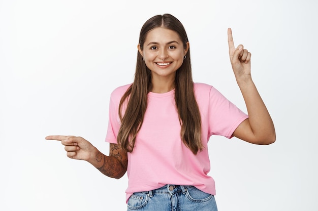 幸せな若い女性の肖像画笑顔、指の両側を指して、白い背景に立って、左と上方向を示しています。