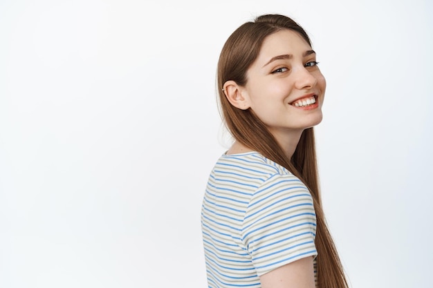 Portrait of happy young woman standing in profile, turn head at camera, looking with joyful smile, white teeth, standing against white background.