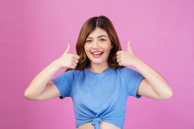 Портрет счастливой молодой женщины в повседневной футболке, показывающей большой палец вверх на розовом фоне