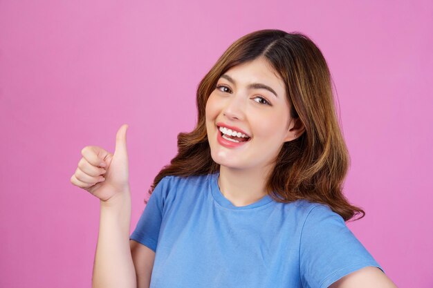 분홍색 배경 위에 고립된 엄지손가락을 보여주는 캐주얼 티셔츠를 입은 행복한 젊은 여성의 초상화