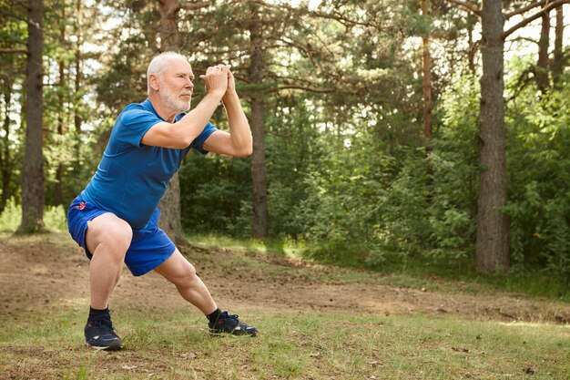 Portrait of healthy active elderly male pensioner in running shoes exercising outdoors, holding hands together in front of him and doing side lunges, having focused concentrated facial expression