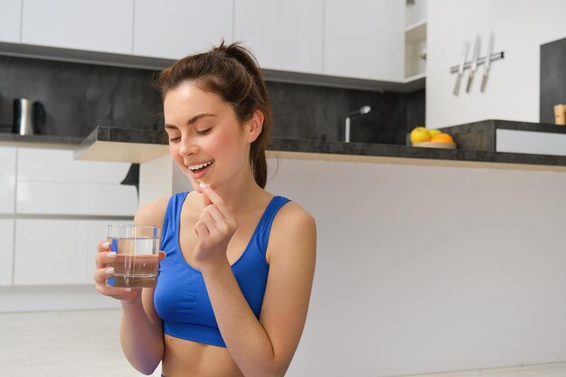 Portrait of healthy smiling young woman taking vitamins after workout holding glass of water and