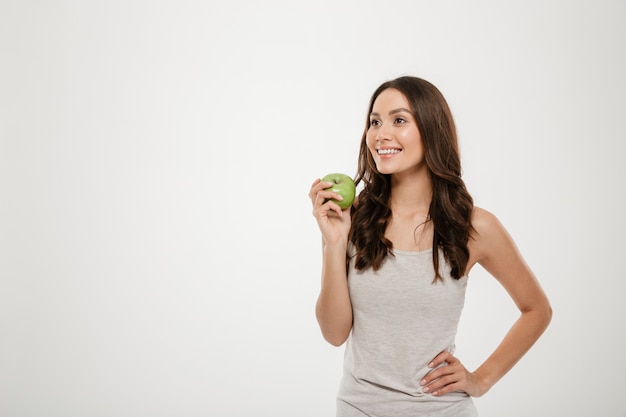 白で隔離された長い茶色の髪立っている健康な女性の肖像画、緑のジューシーなリンゴを試飲