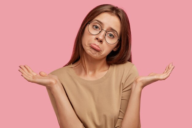 Portrait of hesitant European female purses lips, clasps hands in bewilderment, being uncertain and puzzled, has long hair, wears round spectacles and beige sweater, isolated over pink wall.