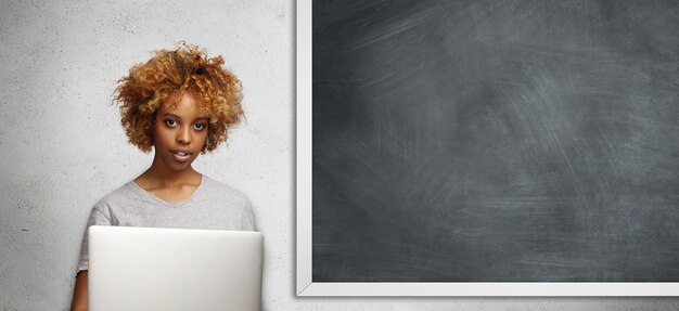 Портрет хипстерского африканского студента с вьющимися волосами и пирсингом на лице, работающего над исследовательским проектом с использованием портативного компьютера, стоя на пустой доске в классе.
