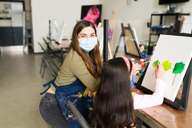 Ritratto di un'insegnante di sesso femminile ispanico con una maschera facciale che insegna a una bambina come dipingere con un pennello durante una lezione di pittura