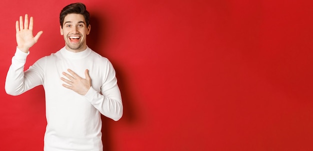 Portrait of honest smiling man in white sweater making a promise swearing to tell truth standing against red background