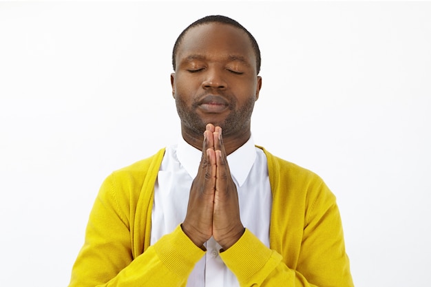 Portrait of hopeful adult african american male closing eyes and pressing palms together, praying, hoping for the best. calm peaceful dark skinned man holding hands in namaste while meditating