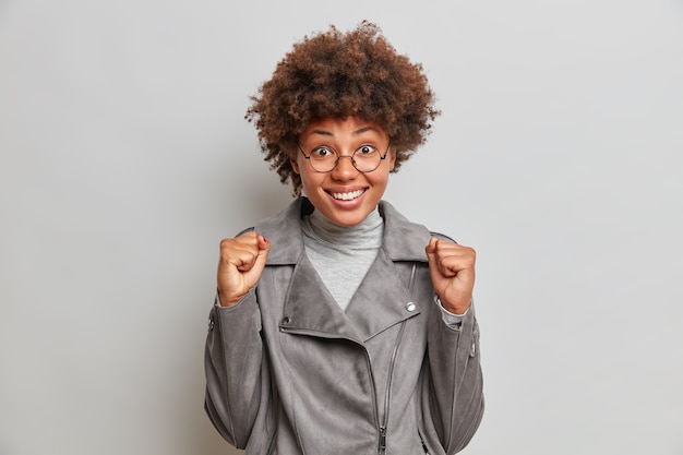 Portrait of hopeful optimistic woman clenches fists, smiles broadly, celebrates success or triumph, wears fashionable grey jacket, stands indoor