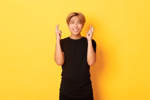 Портрет обнадеживающего взволнованного азиатского парня, скрестив пальцы на удачу, загадывающего желание или ожидание, стоящего на желтой стене