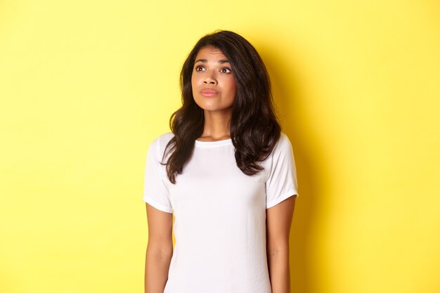 Portrait of hopeful young african-american girl, longing for something, looking at upper left corner dreamy, standing over yellow background.