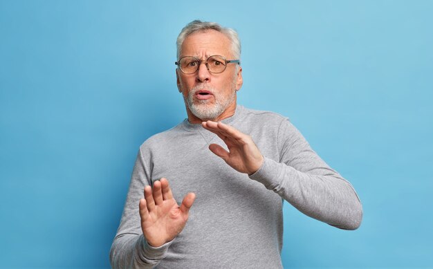 Portrait of horrifiied mature man with grey hair and beard makes frightened gesture tries to defense himself wears transparent glasses and casual sweater covers himself from aggression