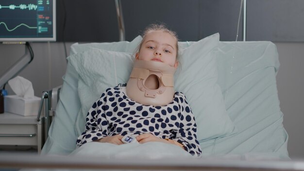 병원 병동에서 의료 상담을 하는 동안 의료 목 경추 칼라를 착용하고 침대에서 쉬고 있는 입원한 어린 아이의 초상화. 고통스러운 사고를 겪고 장애 아이