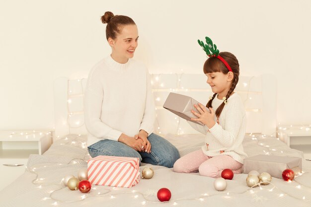 Портрет, если мать и дочь в белых свитерах повседневного стиля, сидя на кровати, ребенок женского пола держит подарочную коробку от мамы, празднует зимние праздники.