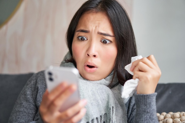 Portrait of ill woman looking at smartphone looking concerned and shocked at mobile phone reading bad news while sitting on sick leave at home