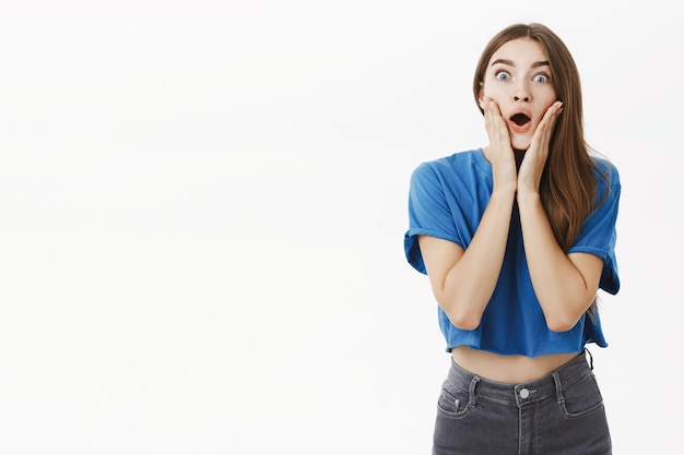 Portrait of impressed and astonished gossip girl with brown hair in blue t-shirt gasping dropping jaw from amazement and surprise touching cheeks popping eyes impressed by rumor