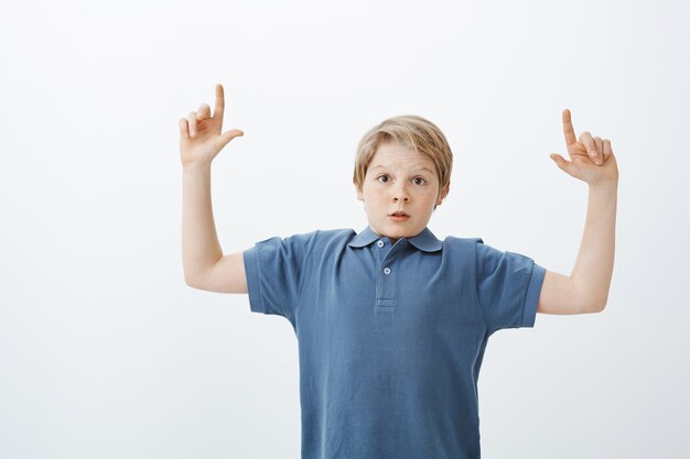 Portrait of impressed excited young european boy with blond hair, raising index fingers and pointing up while being surprised and amazed