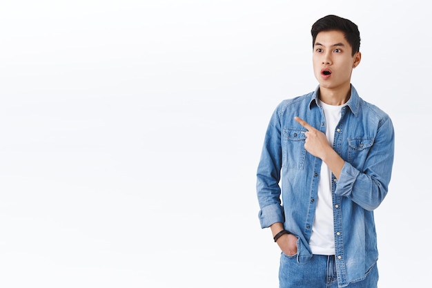 Portrait of impressed, fascinated asian guy seeing something super cool, drop jaw, gasping looking and pointing finger left at awesome discount offer