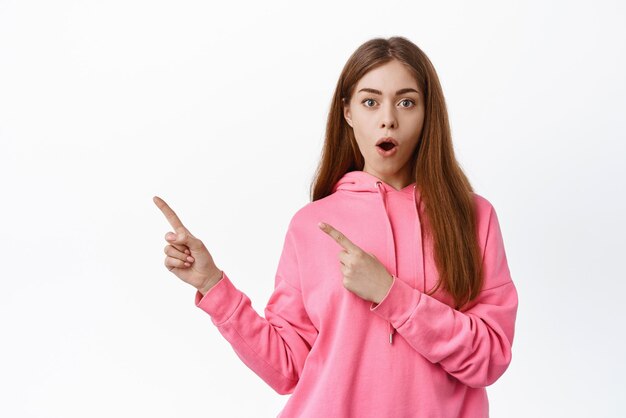 Portrait of impressed girl points left looks with dropped jaw and amazed face checking out advertisement white background